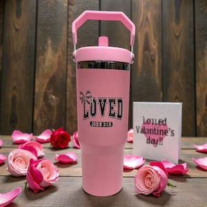 May include: A pink insulated tumbler with a handle, featuring the word "LOVED" and the text "JOHN 3:16" in black. The tumbler is surrounded by pink rose petals and roses. A small card with text is in the background.