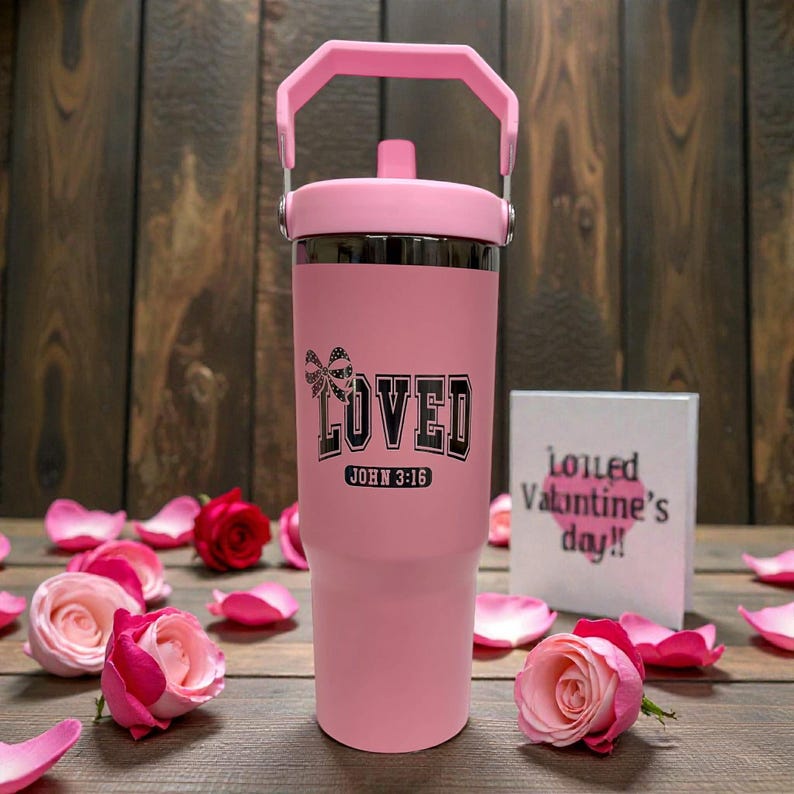 May include: A pink insulated tumbler with a handle, featuring the word "LOVED" and the text "JOHN 3:16" in black. The tumbler is surrounded by pink rose petals and roses. A small card with text is in the background.