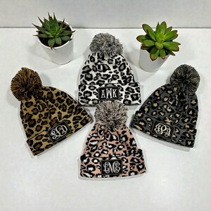 May include: Five knit beanies with pom-poms, each featuring a leopard print pattern in various colors: brown, black and white, gray, and pink. Each beanie has a rectangular patch with a monogram.