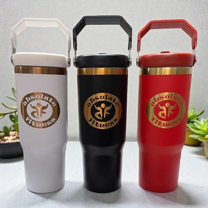 May include: Three insulated tumblers in white, black, and red, each with a gold band and a handle. The tumblers feature the "absolute Fitness" logo in gold. The white tumbler has a white handle, the black has a black handle, and the red has a red handle.
