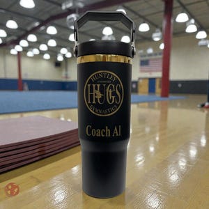 May include: Black tumbler with a black handle and lid, featuring gold accents. The tumbler has the text "HUNTLEY UNLIMITED HUGS GYMNASTICS Coach Al" in gold lettering. The tumbler is on a wooden floor.
