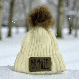 May include: Cream-colored knit beanie with a brown faux fur pom-pom. The hat features a rectangular patch with the name "BRYNLEE" in dark brown lettering. The hat is ribbed and appears to be made of a soft, warm material, perfect for cold weather.