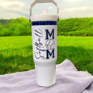 May include: White insulated tumbler with a white handle and lid. The tumbler has a blue band near the top and features the words "Softball Mom" in navy blue, along with a baseball graphic with a heart.