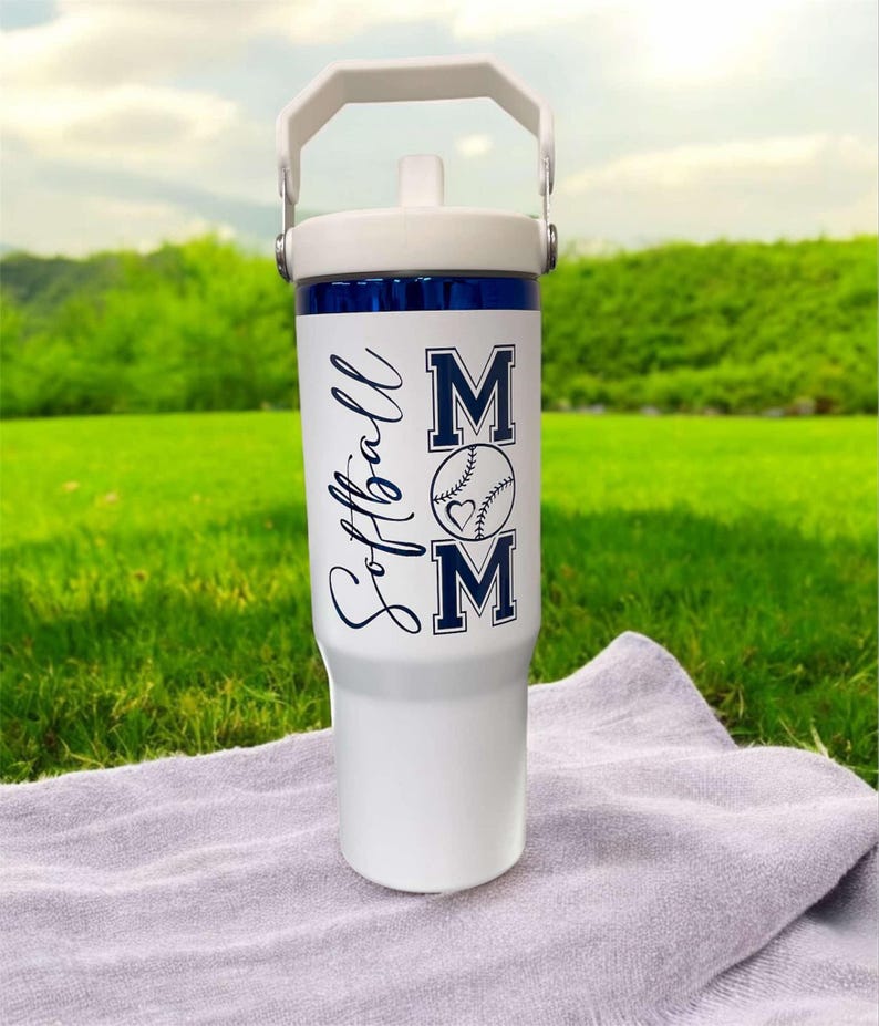 May include: White insulated tumbler with a white handle and lid. The tumbler has a blue band near the top and features the words "Softball Mom" in navy blue, along with a baseball graphic with a heart.