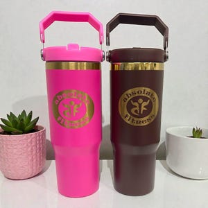 May include: Two insulated tumblers, one bright pink and one dark brown, each with a handle and a gold band. The pink tumbler has a pink handle and the brown tumbler has a brown handle. Both tumblers have the text "Absolute Fitness" in gold.