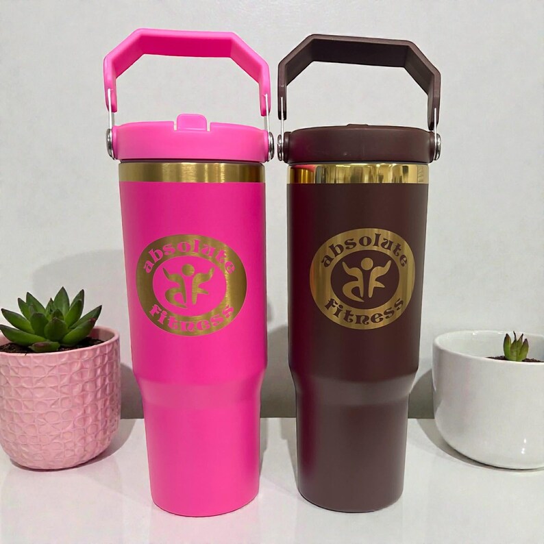 May include: Two insulated tumblers, one bright pink and one dark brown, each with a handle and a gold band. The pink tumbler has a pink handle and the brown tumbler has a brown handle. Both tumblers have the text "Absolute Fitness" in gold.