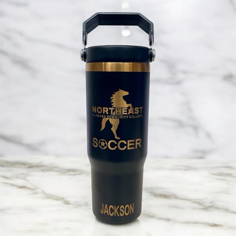 May include: A black insulated tumbler with a gold band and handle. The tumbler features the text "NORTHEAST ALABAMA COMMUNITY COLLEGE" and a gold horse graphic. Below the graphic is the word "SOCCER" and the name "JACKSON".