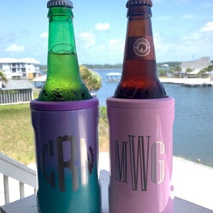 May include: Two insulated beverage holders, one purple and teal, the other pink, each holding a bottle. The purple and teal holder has the initials "CRN" and the pink holder has the initials "MWG". The background shows a body of water and blue sky.