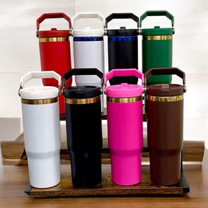 May include: A collection of insulated tumblers in various colors, including red, white, black, green, pink, and brown. Each tumbler has a handle, a lid, and a gold-colored band. The tumblers are displayed on a wooden rack.