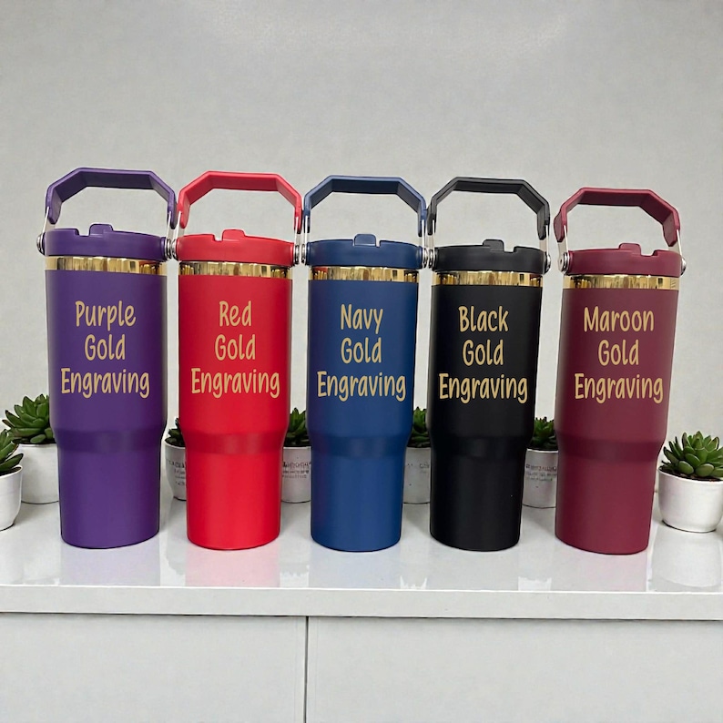 May include: Five insulated tumblers in purple, red, navy, black, and maroon. Each tumbler has a gold rim and handle, with gold engraved text. The text on each tumbler reads "Purple Gold Engraving", "Red Gold Engraving", "Navy Gold Engraving", "Black Gold Engraving", and "Maroon Gold Engraving".