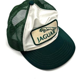 May include: A green and white trucker hat featuring a mesh back and a solid front panel. A rectangular patch is centered on the front, displaying an embroidered green jaguar silhouette above the text JAGUAR in green capital letters.