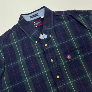 May include: A blue, green, and red plaid button-up shirt with a gold button closure and a small embroidered logo on the left chest pocket. The shirt is made of pure cotton.