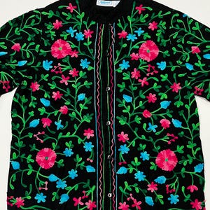 Op de afbeelding: Zwarte jas met kleurrijke bloemenborduurwerk. De jas is versierd met roze, blauwe en groene bloemenborduurwerk. Het borduurwerk is gedetailleerd en bedekt de hele jas, waardoor een kleurrijk en opvallend ontwerp ontstaat.
