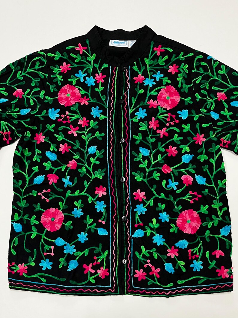 Op de afbeelding: Zwarte jas met kleurrijke bloemenborduurwerk. De jas is versierd met roze, blauwe en groene bloemenborduurwerk. Het borduurwerk is gedetailleerd en bedekt de hele jas, waardoor een kleurrijk en opvallend ontwerp ontstaat.