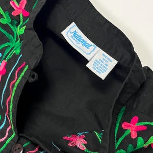 Op de afbeelding: Zwart overhemd met kleurrijke bloemenborduurwerk. De borduursels zijn roze, groen en blauw. Het overhemd heeft een kraag en een label met "National since 1952".