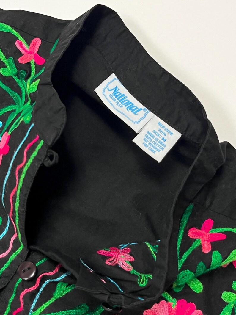 Op de afbeelding: Zwart overhemd met kleurrijke bloemenborduurwerk. De borduursels zijn roze, groen en blauw. Het overhemd heeft een kraag en een label met "National since 1952".