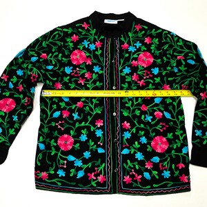Op de afbeelding: Zwarte geborduurde jas met een bloemenmotief in roze, blauw en groen. De jas heeft een knoopsluiting aan de voorkant en lange mouwen. De jas meet ongeveer 51 cm over de borst.