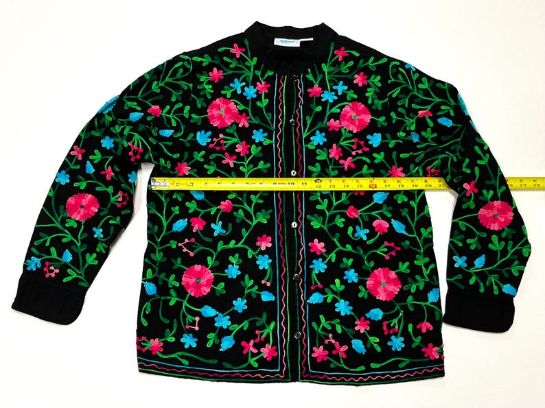 Op de afbeelding: Zwarte geborduurde jas met een bloemenmotief in roze, blauw en groen. De jas heeft een knoopsluiting aan de voorkant en lange mouwen. De jas meet ongeveer 51 cm over de borst.