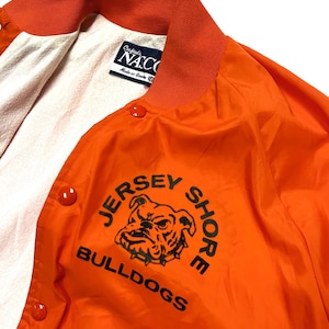 May include: Bright orange jacket with a snap front closure and a contrasting orange collar. The jacket features the text "JERSEY SHORE BULLDOGS" with a bulldog graphic in black. The interior lining is white. The tag reads "Crafted by NATCO Made in America."