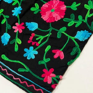 Op de afbeelding: Zwarte stof met levendige bloemenborduurwerk. Het ontwerp toont roze, blauwe en groene geborduurde bloemen en bladeren. De randen zijn afgewerkt met een kleurrijke, golvende steek.