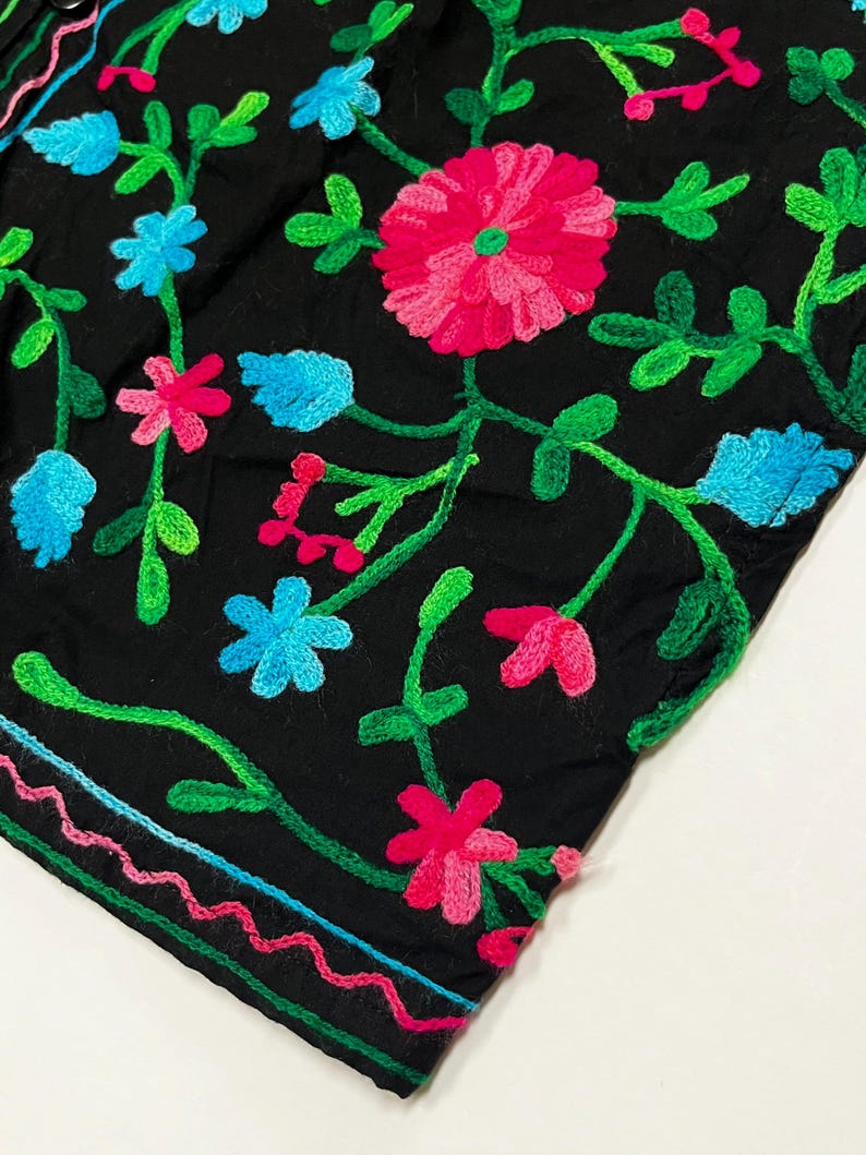 Op de afbeelding: Zwarte stof met levendige bloemenborduurwerk. Het ontwerp toont roze, blauwe en groene geborduurde bloemen en bladeren. De randen zijn afgewerkt met een kleurrijke, golvende steek.