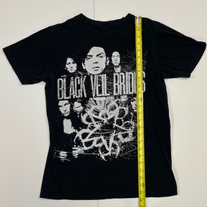 May include: Black t-shirt featuring the band name "Black Veil Brides" in distressed white lettering. The shirt displays a graphic of the band members and abstract white designs. The shirt measures approximately 28 inches in length.