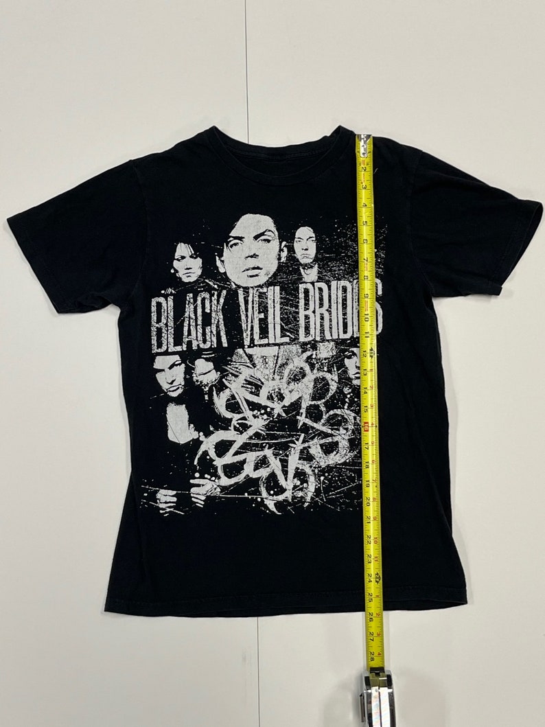 May include: Black t-shirt featuring the band name "Black Veil Brides" in distressed white lettering. The shirt displays a graphic of the band members and abstract white designs. The shirt measures approximately 28 inches in length.