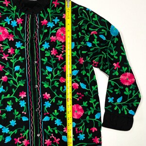 Op de afbeelding: Zwart overhemd met borduursel van roze, blauwe en groene bloemen. Het shirt met lange mouwen heeft een opstaande kraag en is ongeveer 69 cm lang.