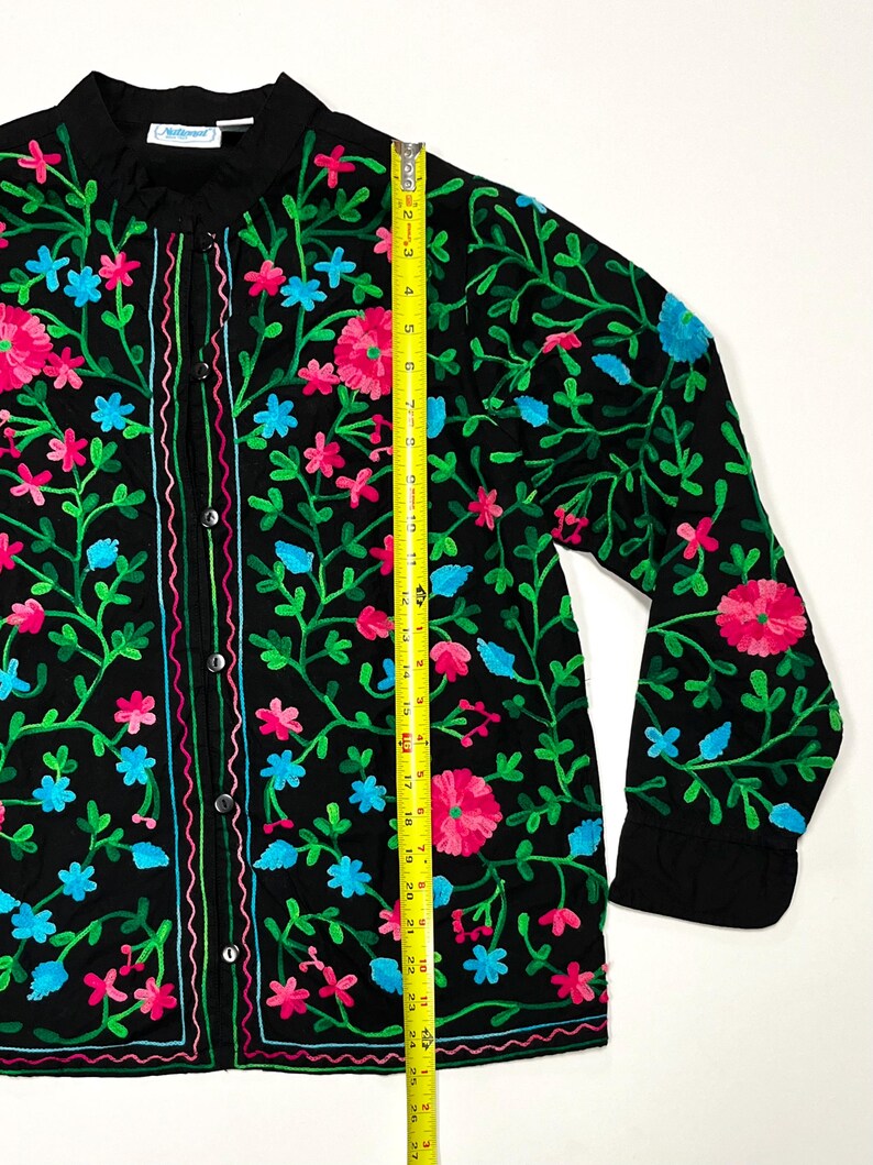 Op de afbeelding: Zwart overhemd met borduursel van roze, blauwe en groene bloemen. Het shirt met lange mouwen heeft een opstaande kraag en is ongeveer 69 cm lang.