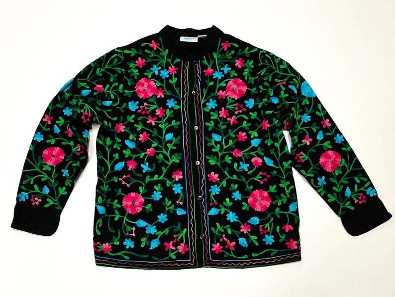 Op de afbeelding: Zwarte geborduurde jas met een bloemenmotief. De jas heeft roze en blauwe bloemen met groene wijnranken en bladeren. De jas heeft een knoopsluiting en lange mouwen.