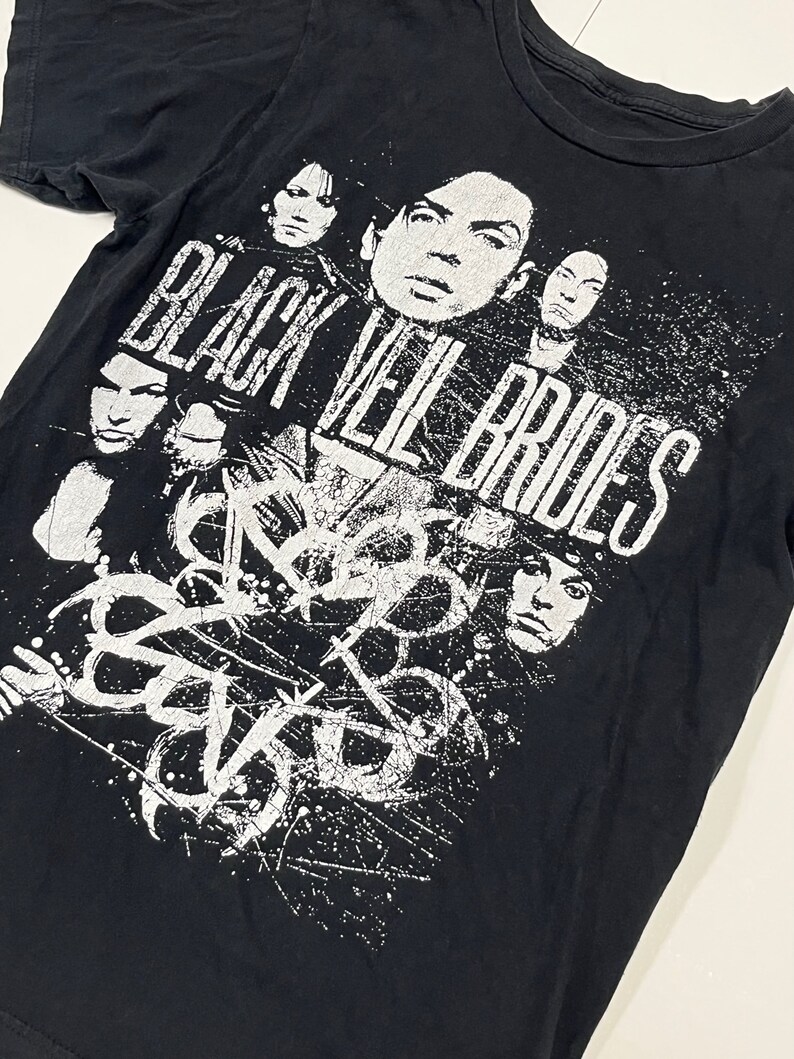 May include: Black t-shirt featuring a distressed white graphic of the band Black Veil Brides. The design includes the band's name and faces of the band members. The shirt is made of a soft, dark fabric.