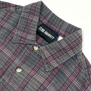May include: Close-up of a gray plaid button-down shirt with burgundy and white accents. The shirt has a collar and buttons down the front. The tag reads "ZERO GRAVITY".