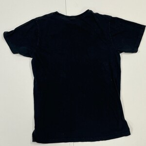 May include: A black short-sleeved t-shirt, laid flat on a white surface. The shirt is plain, with a crew neck and a slightly worn appearance. The fabric appears to be cotton or a similar material. The shirt is a basic, everyday garment.