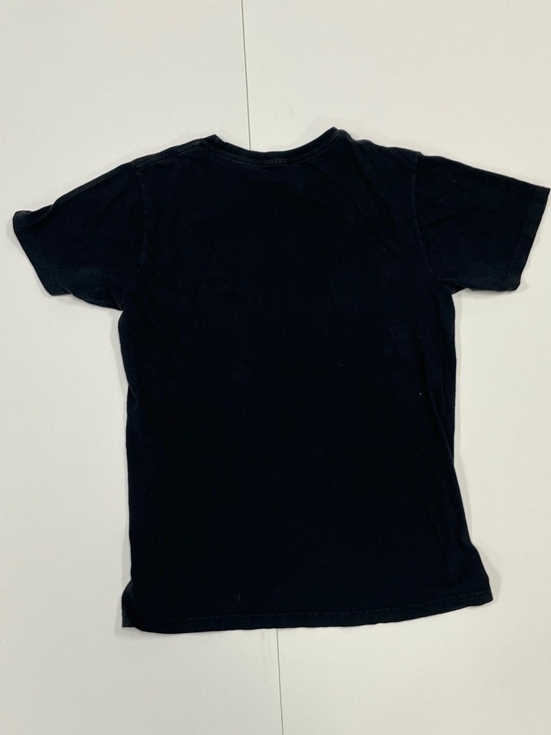 May include: A black short-sleeved t-shirt, laid flat on a white surface. The shirt is plain, with a crew neck and a slightly worn appearance. The fabric appears to be cotton or a similar material. The shirt is a basic, everyday garment.