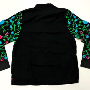 Op de afbeelding: Zwart shirt met lange mouwen en bloemenborduurwerk op de mouwen. Het borduurwerk heeft groene bladeren, roze en blauwe bloemen. Het shirt heeft een mandarijnkraag en manchetten met knopen. Het shirt is gemaakt van een zacht, comfortabel materiaal.