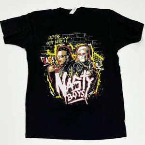 May include: Black t-shirt featuring a graphic of two men with sunglasses, the words "NASTY BOYS" in a graffiti-style font, and the phrases "Let's Get Nasty", "Sags", and "Knobs".