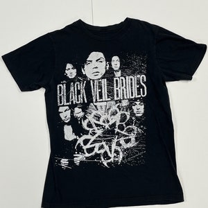 May include: Black t-shirt featuring a white graphic of a band with the text "BLACK VEIL BRIDES". The design includes portraits of band members and abstract shapes. The shirt is made of a soft material.