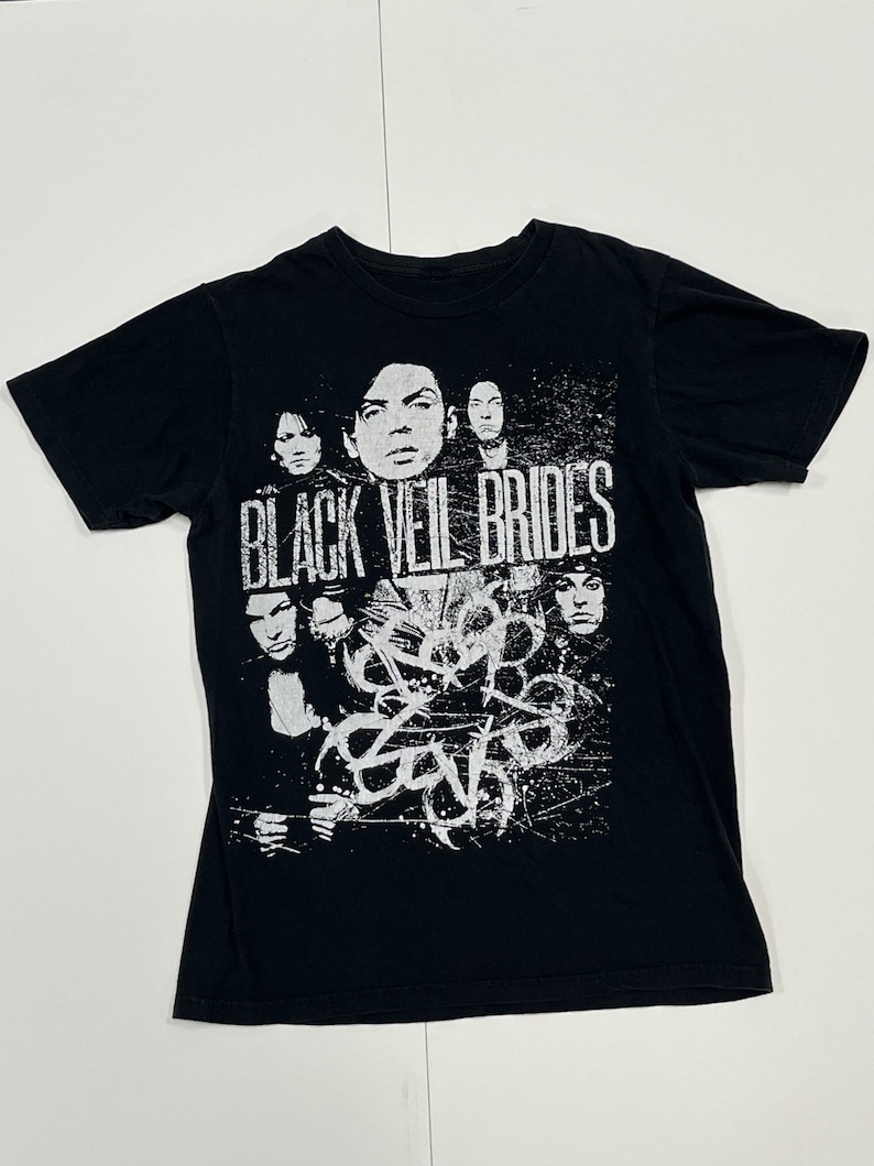May include: Black t-shirt featuring a white graphic of a band with the text "BLACK VEIL BRIDES". The design includes portraits of band members and abstract shapes. The shirt is made of a soft material.