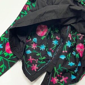 Op de afbeelding: Zwart kledingstuk met levendige bloemenborduurwerk in roze, blauw en groen. Het borduurwerk heeft gedetailleerde bloemen- en bladmotieven. Het kledingstuk lijkt van een zachte stof te zijn.