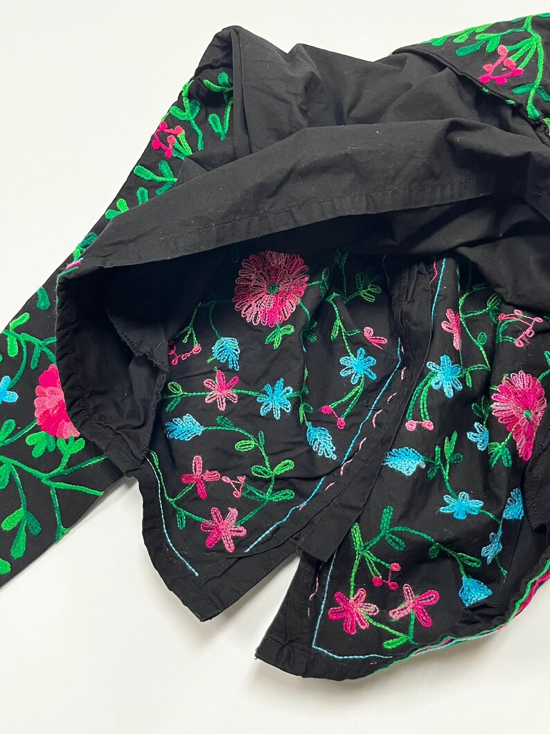 Op de afbeelding: Zwart kledingstuk met levendige bloemenborduurwerk in roze, blauw en groen. Het borduurwerk heeft gedetailleerde bloemen- en bladmotieven. Het kledingstuk lijkt van een zachte stof te zijn.