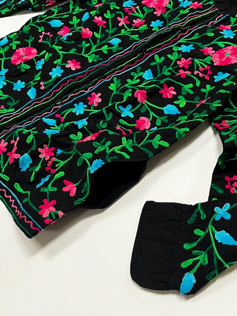 Op de afbeelding: Zwart overhemd met gedetailleerde bloemenborduurwerk. Het borduurwerk toont roze, blauwe en groene bloemen en ranken. Het overhemd heeft lange mouwen en een klassieke kraag. Het ontwerp is gedetailleerd en levendig.