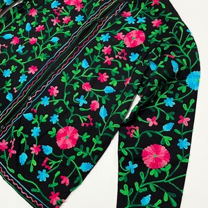 Op de afbeelding: Zwart overhemd met bloemenborduurwerk in roze, blauw en groen. Het shirt met lange mouwen heeft een opstaande kraag en een knoopsluiting. Het kledingstuk is versierd met geborduurde bloemen en ranken.