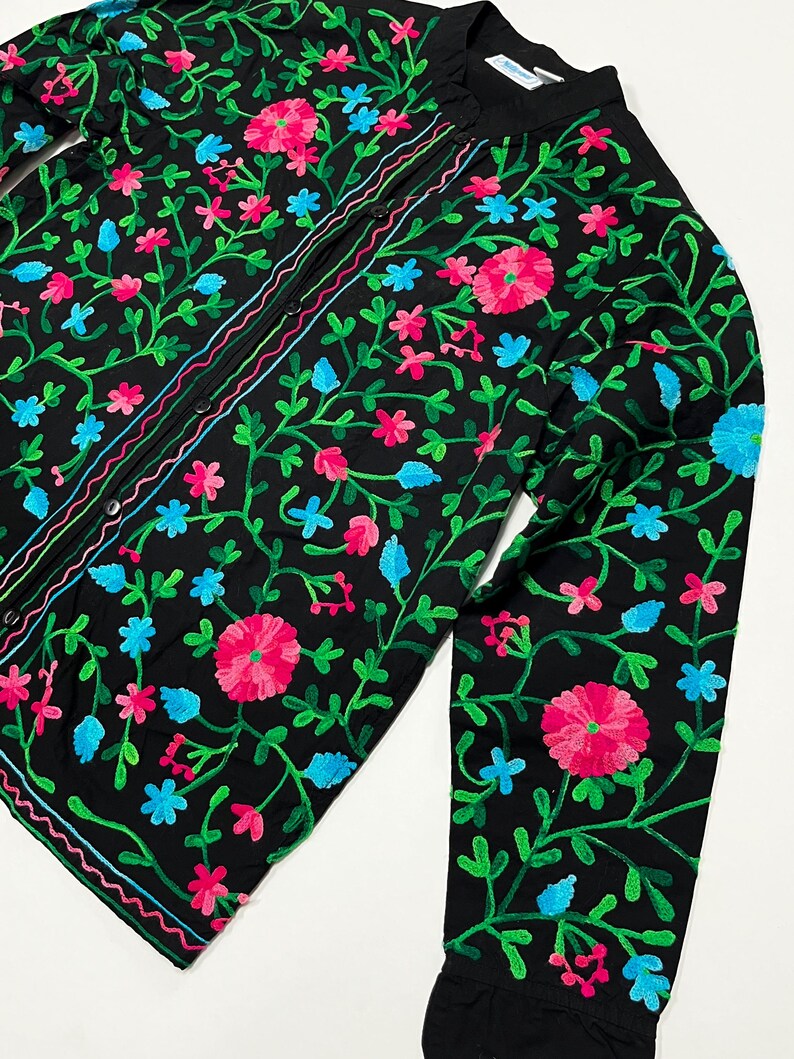 Op de afbeelding: Zwart overhemd met bloemenborduurwerk in roze, blauw en groen. Het shirt met lange mouwen heeft een opstaande kraag en een knoopsluiting. Het kledingstuk is versierd met geborduurde bloemen en ranken.