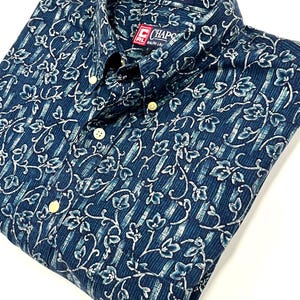 May include: A blue button-down shirt with a floral pattern and vertical stripes. The shirt has a classic collar and the brand name "Chaps" is visible. Made of 100% cotton.