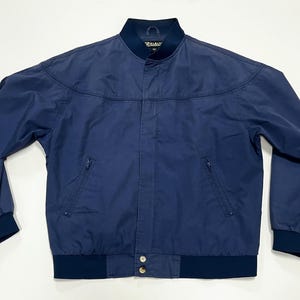 May include: A navy blue bomber jacket with a zipper closure and a ribbed collar and cuffs. The jacket has two zippered pockets on the front and a button closure at the bottom.