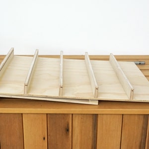 May include: A wooden shelf unit with five slots, made of light-colored plywood. The shelf is angled slightly and is sitting on a wooden surface.