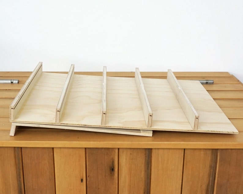 May include: A wooden shelf unit with five slots, made of light-colored plywood. The shelf is angled slightly and is sitting on a wooden surface.