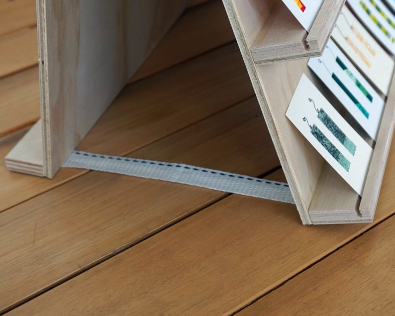 May include: A wooden display stand with multiple shelves, each holding a card with a green and white design. The stand is made of light wood and has a white strap attached to the bottom.