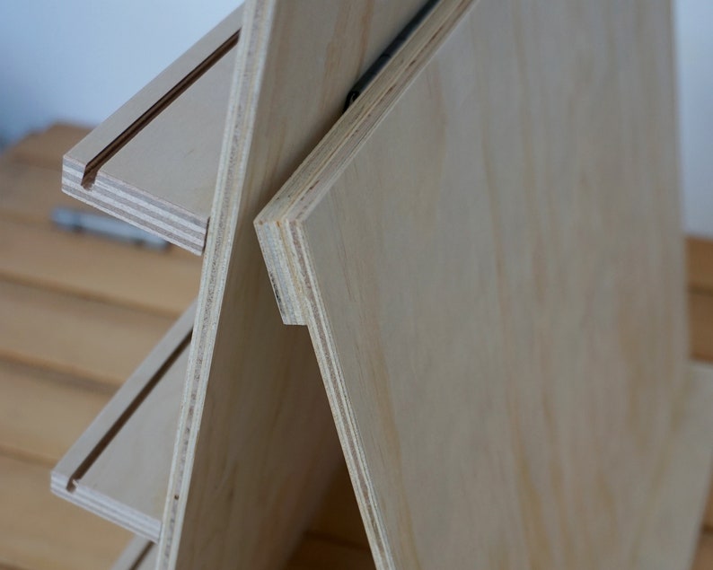 May include: A wooden tiered stand with three shelves. The stand is made of light-colored wood and has a natural finish. The shelves are slightly angled and have a groove on the front edge.