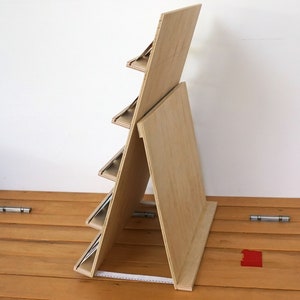 May include: A wooden display stand with five shelves for showcasing items. The stand is made of light brown plywood and has a simple, modern design.